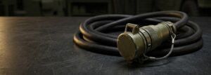 Heavy-duty military slave cable assembly with a capped, ruggedized plug connector coiled on a steel workbench in a military vehicle maintenance bay